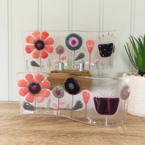May include: Two glass panels with a wavy design feature a colourful floral pattern. The panels are stacked on top of each other and have a wooden base. The flowers are pink, red, purple, and grey.