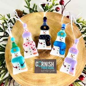 May include: Five handmade glass snowman ornaments with different coloured hats, scarves, and accents. The ornaments are hanging from white ribbon on a wooden surface. The text "CORNISH FUSED GLASS by Kirsty Smith" is visible on a card below the ornaments.