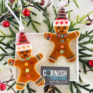 May include: Two gingerbread man Christmas ornaments made of fused glass. Each ornament has a brown body, a red and white hat, and colorful details. The ornaments are displayed in a white box with a sprig of greenery and red berries.