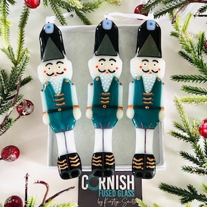 Könnte beinhalten: Drei tealfarbene und weiße Nussknacker-Anhänger aus Glas mit goldenen Akzenten. Die Anhänger hängen an einer weißen Schnur und werden in einer weißen Schachtel mit dem Text "CORNISH FUSED GLASS by Kirsty Smith" präsentiert.