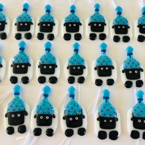 Puede incluir: Una colecci&oacute;n de adornos de ovejas blancas con sombreros de fiesta azules y dorados. Cada oveja tiene ojos negros y una nariz negra. Los adornos cuelgan de cintas blancas.