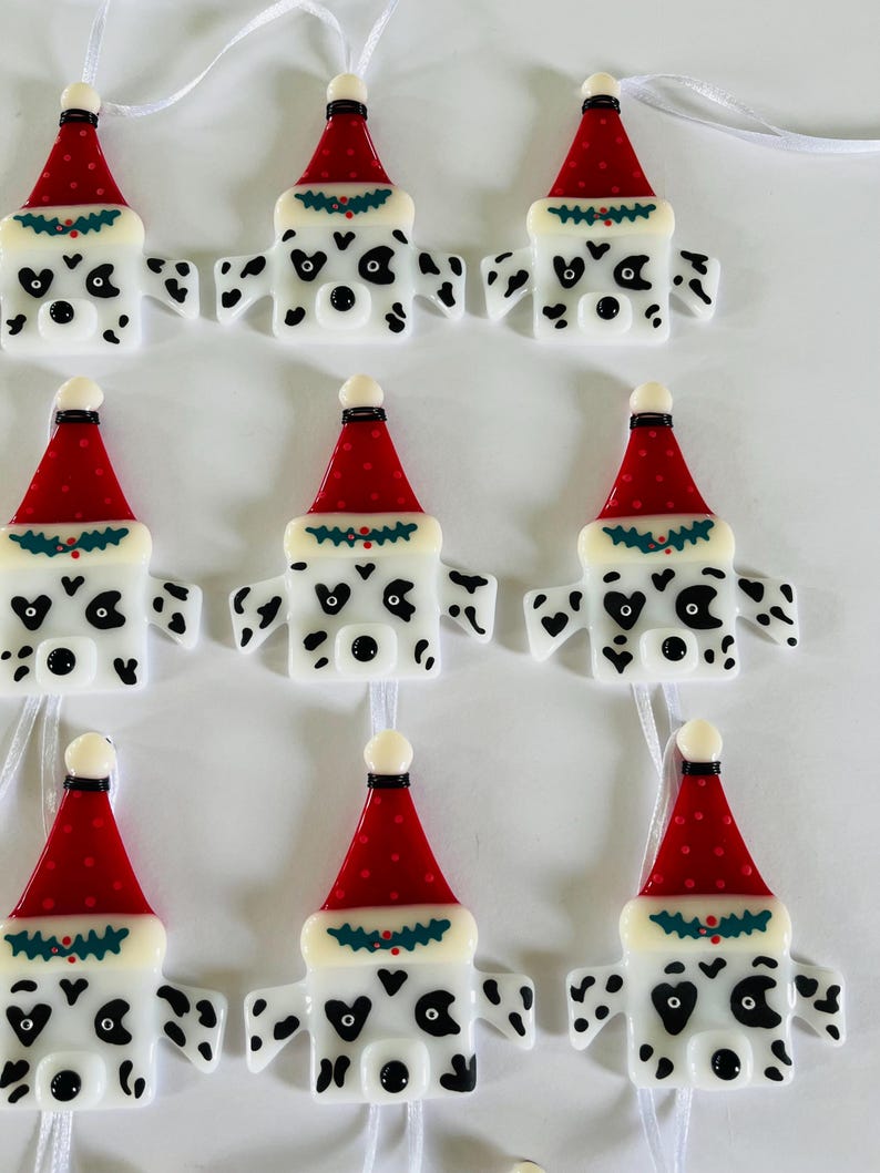 May include: Eight white and black Dalmatian dog ornaments with red and green Santa hats. The ornaments are made of glass and have a white background with black spots. Each ornament has a loop for hanging.