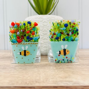 May include: Two small glass candle holders shaped like flower pots with a bee design. The pots are painted a light blue colour with white polka dots and a gold rim. The flowers are made of colourful glass pieces.