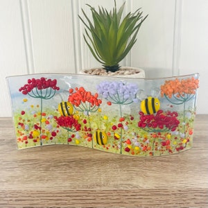 May include: A wavy glass art piece with a floral design featuring red, orange, and purple flowers with green stems and leaves. Three yellow and black bees are flying around the flowers. The background is a light blue with white dots.