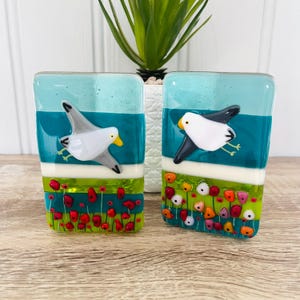 May include: Two glass art pieces with a seagull design. Each piece features a seagull against a teal and blue background, with a field of red and colorful flowers at the bottom. The pieces are rectangular and have a glossy finish.
