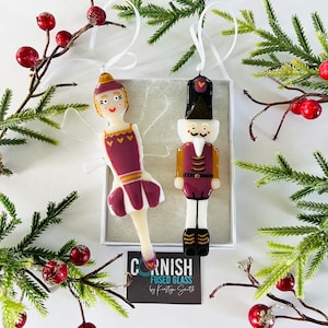 Puede incluir: Un conjunto de dos adornos de vidrio, una bailarina y un cascanueces, en una caja blanca con una etiqueta dorada y negra que dice "Cornish Fused Glass by Kristyn Smith". La bailarina lleva un vestido burdeos y una corona dorada. El cascanueces lleva un uniforme burdeos con detalles dorados. Ambos adornos están decorados con detalles dorados.