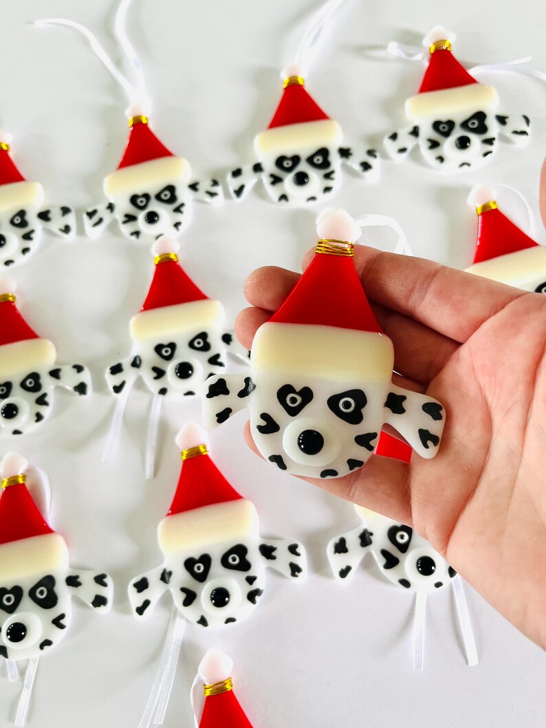 May include: A set of handmade glass ornaments featuring Dalmatian dog faces wearing Santa hats. The ornaments are white with black spots and have black eyes and noses. Each ornament has a white ribbon for hanging.