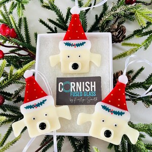 May include: Three white glass ornaments shaped like dogs wearing red Santa hats with white polka dots and green holly. The ornaments are handmade and say "CORNISH FUSED GLASS by Kirsty Smith" on the box.