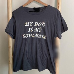 May include: Dark gray t-shirt with the text "MY DOG IS MY SOULMATE" in white letters with floral accents. The shirt is hanging on a wooden hanger against a light background. The shirt has short sleeves.