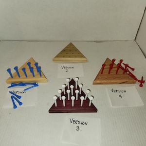 May include: Four versions of a triangular wooden game with pegs. Version 1 has blue pegs, version 2 has wooden pegs, version 3 has white pegs, and version 4 has red pegs. Each version has a label with the version number.
