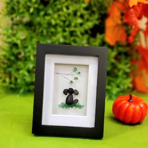 May include: A framed pebble art piece featuring a dog design. The dog is crafted from dark gray pebbles, with green glass pebbles representing grass and leaves. The frame is black, and a small orange pumpkin is in the background.