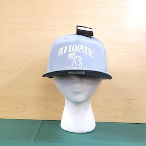 May include: A light blue trucker hat with a black brim and mesh back. The hat features the text "NEW HAMPSHIRE" above an illustration of a moose, with the words "LIVE FREE OR DIE" below. A tag is attached to the top of the hat.