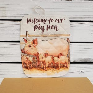 May include: A rectangular quilted oven mitt featuring a printed illustration of a large pig and three piglets standing in a pen. The text "Welcome to our pig pen" is printed in dark brown script at the top. The mitt includes a white hanging loop.
