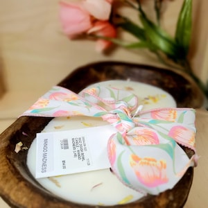 May include: A wooden dough bowl candle with a white wax interior. The candle is tied with a ribbon featuring a floral pattern. The label reads "MANGO MADNESS".