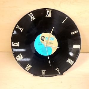 May include: A black vinyl record clock with gold-colored hands and Roman numeral hour markers. The clock face features a blue and gold record label. The clock is displayed on a small black stand.