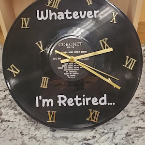 May include: A black vinyl record clock with gold Roman numeral hour markers. The clock face has the text "Whatever, I'm Retired..." printed on it. The record is from the album "Loma Digs Keely Smith" by Coronet Hi-Fi.