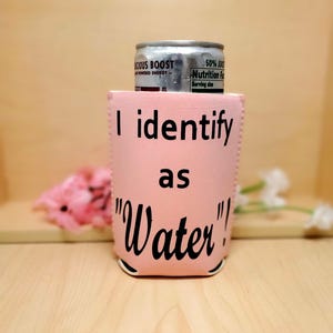 May include: A pink beverage can cooler with the text "I identify as 'Water'!" in black. A silver can of "Delicious Boost" is inside the cooler. The cooler is on a wooden surface.