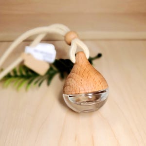 May include: A teardrop-shaped car air freshener with a wooden top and a clear glass bottom. The freshener is suspended by a cream-colored cord and a wooden bead. The background is a light wooden surface.