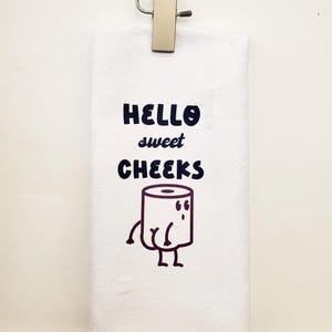 May include: White kitchen towel with the text "Hello sweet cheeks" and a cartoon toilet paper roll with legs and a face.