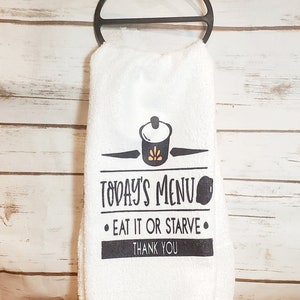 May include: White kitchen towel with black text that reads "Today's Menu - Eat it or Starve - Thank You". A black and gold pot icon is above the text.