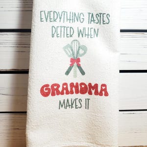 May include: White kitchen towel with the text "Everything tastes better when Grandma makes it" in green and red lettering. A red bow ties together two green kitchen utensils.