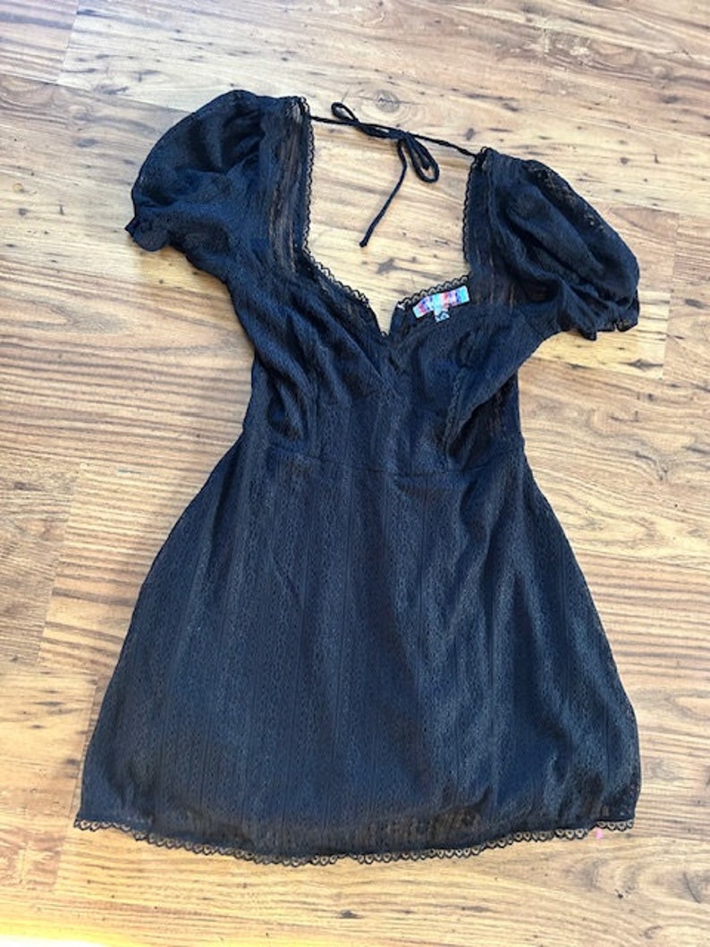 Peut inclure: Une robe en dentelle noire avec un d&eacute;collet&eacute; en c&oelig;ur et des manches bouffantes. La robe a un n&oelig;ud &agrave; l'arri&egrave;re du cou.