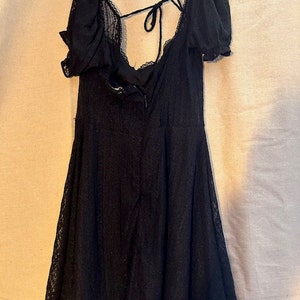 Peut inclure: Une robe courte en dentelle noire avec un d&eacute;collet&eacute; en c&oelig;ur et des manches bouffantes. La robe a un lien &agrave; nouer &agrave; l'arri&egrave;re du cou et un ourlet festonn&eacute;.