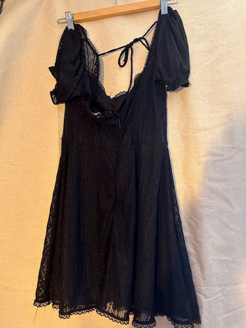 Peut inclure: Une robe courte en dentelle noire avec un d&eacute;collet&eacute; en c&oelig;ur et des manches bouffantes. La robe a un lien &agrave; nouer &agrave; l'arri&egrave;re du cou et un ourlet festonn&eacute;.