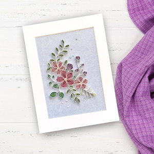 Puede incluir: Un marco blanco con un fondo azul claro que presenta un ramo de flores de papel con flores rosas, rojas y moradas y hojas verdes. Las flores están hechas de papel y están dispuestas en forma de ramo. Hay pequeñas cuentas blancas y azules esparcidas alrededor de las flores.