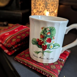 May include: A white ceramic mug with a strawberry plant illustration, resting on a red patterned coaster. The mug has a curved handle and the text "Virginia Meadow Strawberry". A red and white striped fabric is visible.
