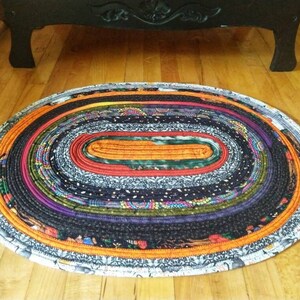 May include: An oval-shaped braided rug with a multi-colored design. The rug is made of fabric scraps and has a white, orange, black, green, purple, and red color scheme.