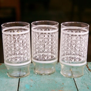 May include: Three vintage clear drinking glasses with a white geometric pattern. The glasses have a cylindrical shape with a slightly flared base and a white band around the top and bottom. The glasses are displayed on a weathered, turquoise-colored wooden surface.
