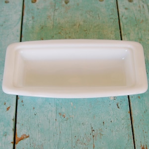 May include: A rectangular, white ceramic dish with rounded edges. The dish is empty and sits on a weathered, turquoise-painted wooden surface. The dish is likely for holding small items.