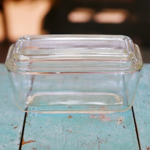 May include: A clear, rectangular glass dish with a matching lid. The dish is sitting on a weathered, light blue wooden surface. The glass appears to be vintage or antique, with a simple, functional design. The dish is likely for food storage.