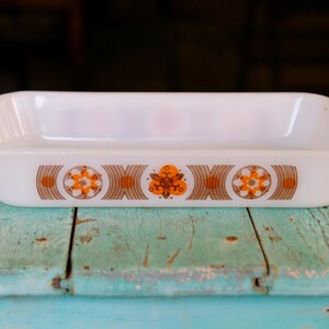 May include: White rectangular baking dish with orange and brown floral design. The dish has rounded corners and handles. The design features a repeating pattern of orange flowers and brown curved lines. The dish is sitting on a weathered, light blue wooden surface.