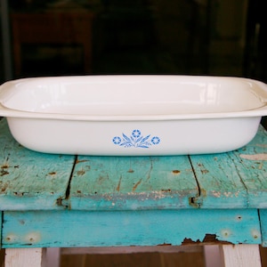 May include: A rectangular white ceramic baking dish with rounded corners and a wide rim. The exterior features a centered blue floral pattern consisting of small flowers and leaves. The dish has a smooth, glossy finish and a deep, open interior.