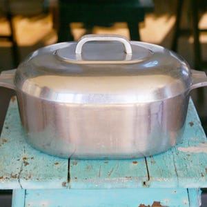 May include: A large, silver-colored cooking pot with a lid and handle, resting on a weathered, turquoise-painted wooden stool. The pot has side handles and a rounded shape, suitable for various cooking tasks.