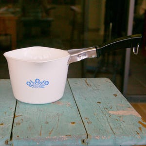 May include: A white ceramic saucepan with a pouring spout and a black handle featuring a silver metal attachment. The exterior of the pan is decorated with a blue floral pattern consisting of small flowers and leaves centered on the side.