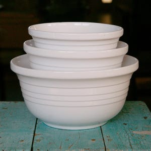 May include: A stack of three white ceramic mixing bowls of varying sizes. Each bowl has a rounded rim and horizontal gray lines around the exterior. The bowls are on a weathered, turquoise-painted wooden surface.