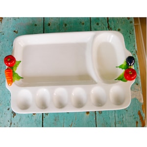 May include: A white ceramic serving platter with multiple compartments. The platter has a rectangular section, an oval section, and six egg-shaped indentations. Decorative red tomatoes, carrots, and olives adorn the sides. The platter is on a distressed turquoise surface.