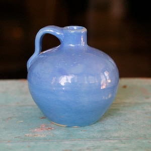 May include: A small, light blue ceramic pitcher with a rounded body and a single handle. The pitcher is sitting on a light blue wooden surface.