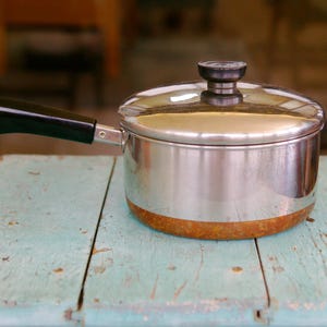 May include: A stainless steel saucepan with a copper-colored base and a black handle. The pot features a matching stainless steel lid with a black knob. A small metal ring is attached to the end of the handle for hanging storage purposes.