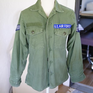 May include: An olive green long-sleeve military shirt featuring a button-down front, two chest pockets, and blue patches on the sleeves. A blue rectangular patch with the white text U.S. AIR FORCE is sewn above the left chest pocket.