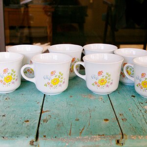 May include: A set of eight white ceramic mugs with a floral design in yellow, orange, and green. Each mug has a handle and is displayed on a weathered, turquoise-painted wooden surface. The mugs are arranged in a semi-circle.