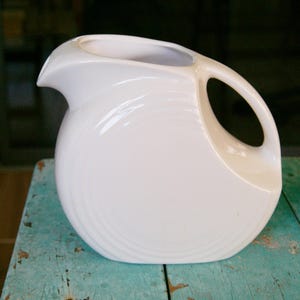 May include: A white ceramic pitcher with a rounded body and a curved handle. The pitcher has a spout for pouring and a capacity of approximately 64 ounces. The design features subtle concentric rings around the base. The pitcher is likely for serving beverages.