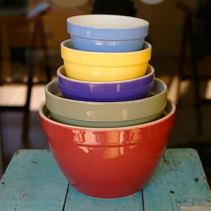 May include: A stack of six ceramic mixing bowls in various colors: red, green, purple, yellow, and blue. The bowls are nested, with the largest red bowl at the bottom and the smallest blue bowl at the top. The bowls are on a weathered blue wooden surface.