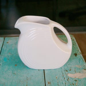 May include: A white ceramic pitcher with a rounded body and a curved handle. The pitcher has a spout for pouring and a series of concentric ridges around the base. The pitcher is sitting on a weathered, turquoise-painted wooden surface.