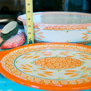 May include: A collection of orange and white ceramic kitchenware, including a serving platter, casserole dish, and serving spoon. The platter is oval with a floral design. The casserole dish has a lid. The spoon is white with an orange floral design. The tape measure shows the casserole dish is about 4 inches tall.