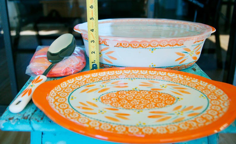 May include: A collection of orange and white ceramic kitchenware, including a serving platter, casserole dish, and serving spoon. The platter is oval with a floral design. The casserole dish has a lid. The spoon is white with an orange floral design. The tape measure shows the casserole dish is about 4 inches tall.