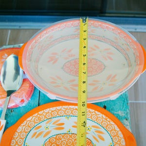 May include: An oval serving dish with a clear lid, orange handles, and a floral design. The dish measures approximately 8 inches in diameter. A matching spoon and trivet are also visible.